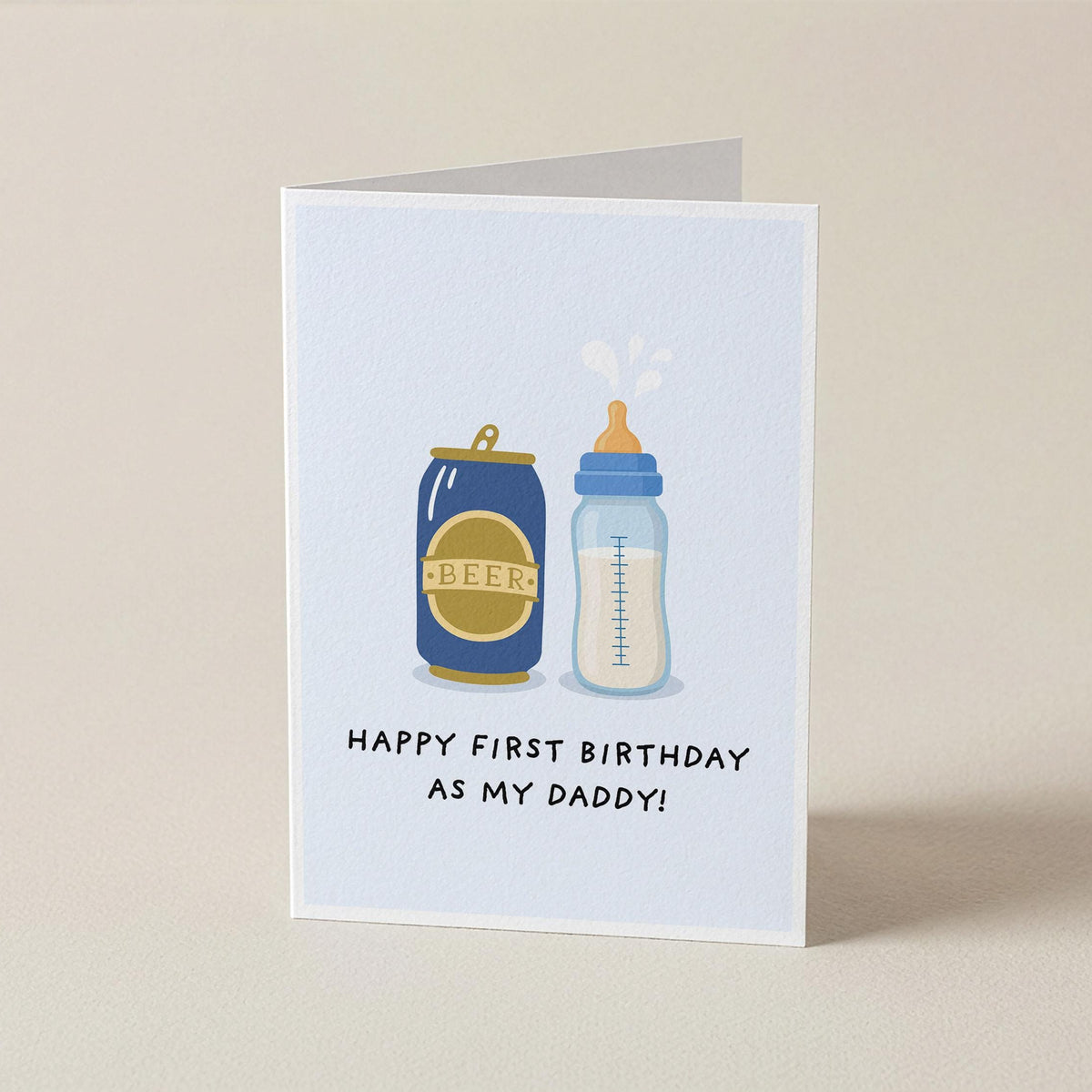 A folded greeting card standing upright on a light beige surface. The card has a white front with a simple illustration of a blue baby bottle and a blue beer can with gold labels, plus the text 'HAPPY FIRST BIRTHDAY AS MY DADDY!'.