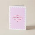 A folded greeting card standing upright on a light beige surface. The card has a white border with a scalloped pink inner design and placeholder red text reading 'YOUR PERSONALISED TEXT HERE' with a small red heart below.