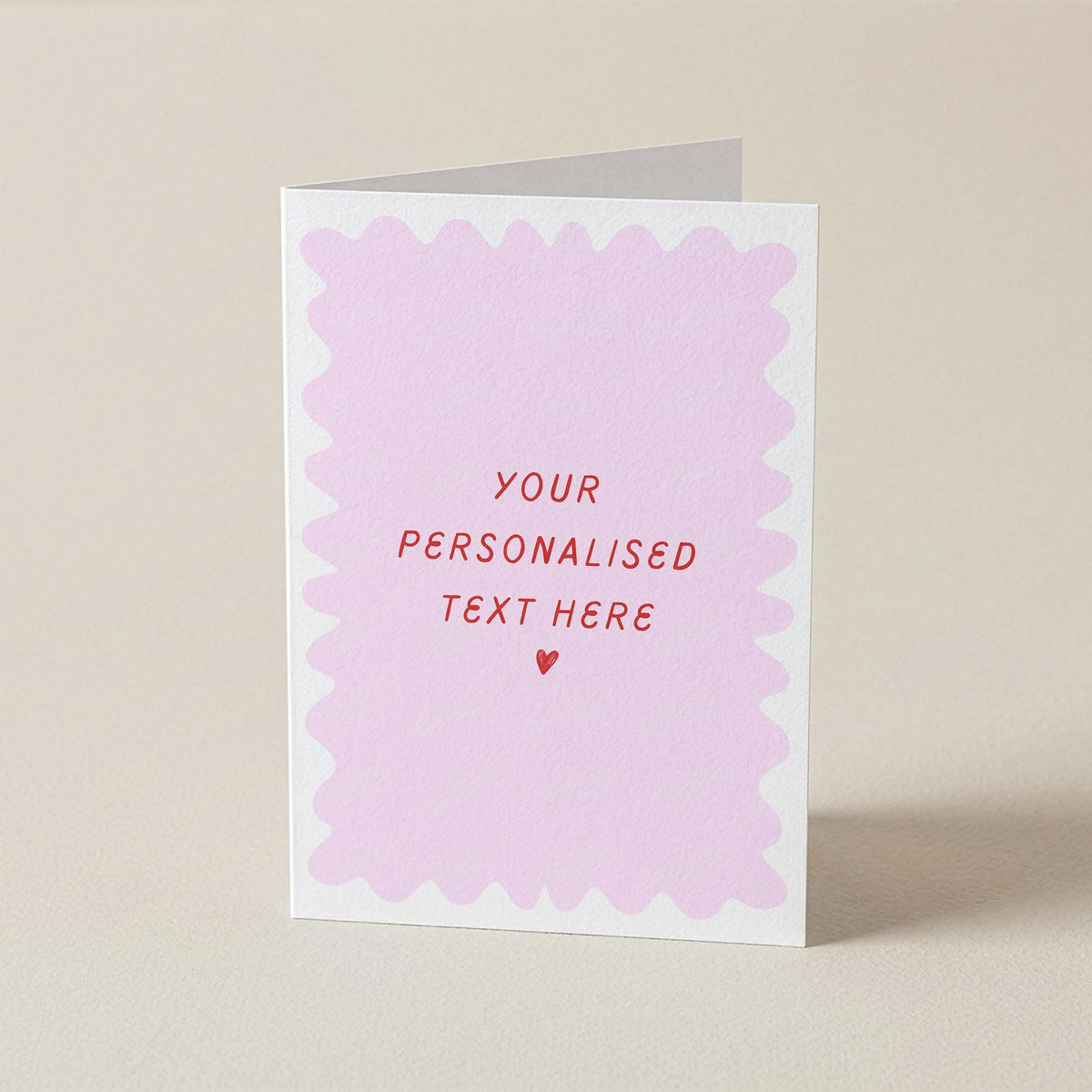 A folded greeting card standing upright on a light beige surface. The card has a white border with a scalloped pink inner design and placeholder red text reading 'YOUR PERSONALISED TEXT HERE' with a small red heart below.