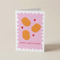A folded greeting card standing upright on a light beige surface. The card has a pink scalloped border with three orange circular kiss marks, small red hearts, and the text 'NUGS AND KISSES' in red at the bottom.