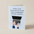 A folded greeting card standing upright on a light beige surface. The card has a pale blue front with the text 'GOOD LUCK CONVINCIING YOUR NEW COWORKERS YOU'RE NORMAL!' above an illustration of hands typing on a laptop.