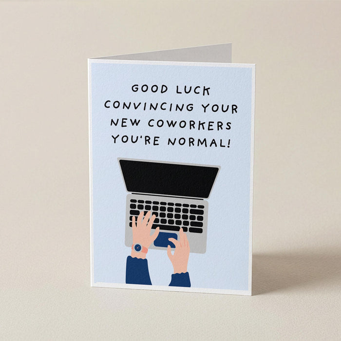 A folded greeting card standing upright on a light beige surface. The card has a pale blue front with the text 'GOOD LUCK CONVINCIING YOUR NEW COWORKERS YOU'RE NORMAL!' above an illustration of hands typing on a laptop.