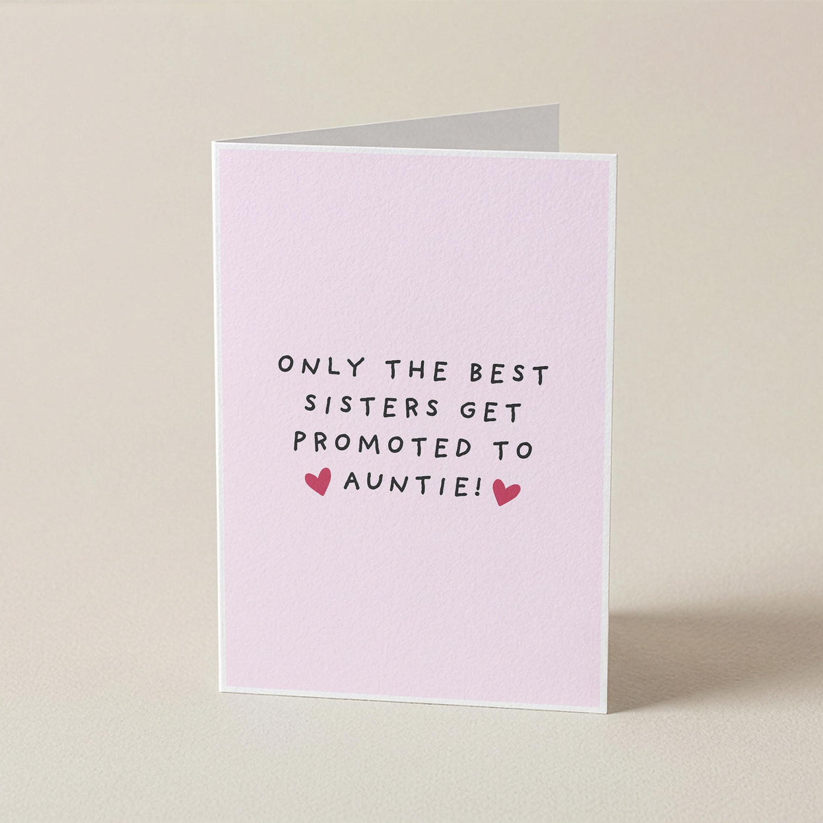 A folded greeting card standing upright on a light beige surface. The card is pale pink with the printed message 'ONLY THE BEST SISTERS GET PROMOTED TO AUNTIE!' in black text and two small pink hearts.