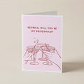 A folded greeting card standing upright on a light beige surface. The card has a pale pink background with red text reading 'GEORGIA, WILL YOU BE MY BRIDESMAID?' and a red line drawing of two clinking cocktail glasses with small sparkles.