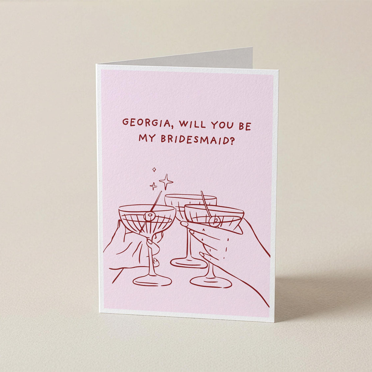 A folded greeting card standing upright on a light beige surface. The card has a pale pink background with red text reading 'GEORGIA, WILL YOU BE MY BRIDESMAID?' and a red line drawing of two clinking cocktail glasses with small sparkles.