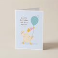 A folded greeting card standing upright on a light beige surface. The card features a pale blue background with a cartoon goose wearing a pink party hat and holding a teal balloon, next to the text 'HAPPY BIRTHDAY YOU SILLY GOOSE!'.