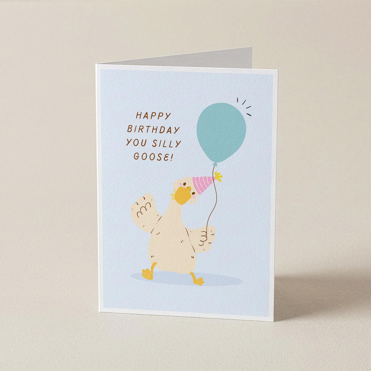 A folded greeting card standing upright on a light beige surface. The card features a pale blue background with a cartoon goose wearing a pink party hat and holding a teal balloon, next to the text 'HAPPY BIRTHDAY YOU SILLY GOOSE!'.