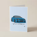 A folded greeting card standing upright on a beige surface. The card has a light blue background with an illustration of a blue sports car and the text 'I got you a birthday car' with 'di' written below.