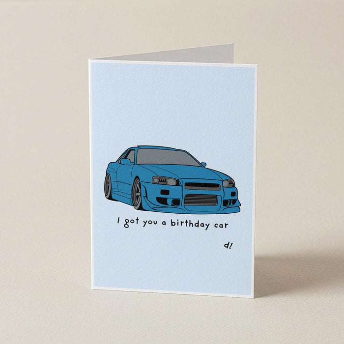 A folded greeting card standing upright on a beige surface. The card has a light blue background with an illustration of a blue sports car and the text 'I got you a birthday car' with 'di' written below.