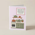 A folded greeting card standing upright on a light beige surface. The card has a pale pink background with a cartoon kitchen illustration and the text, 'I CAN'T WAIT TO CHAT SHIT IN YOUR KITCHEN WITH YOU.