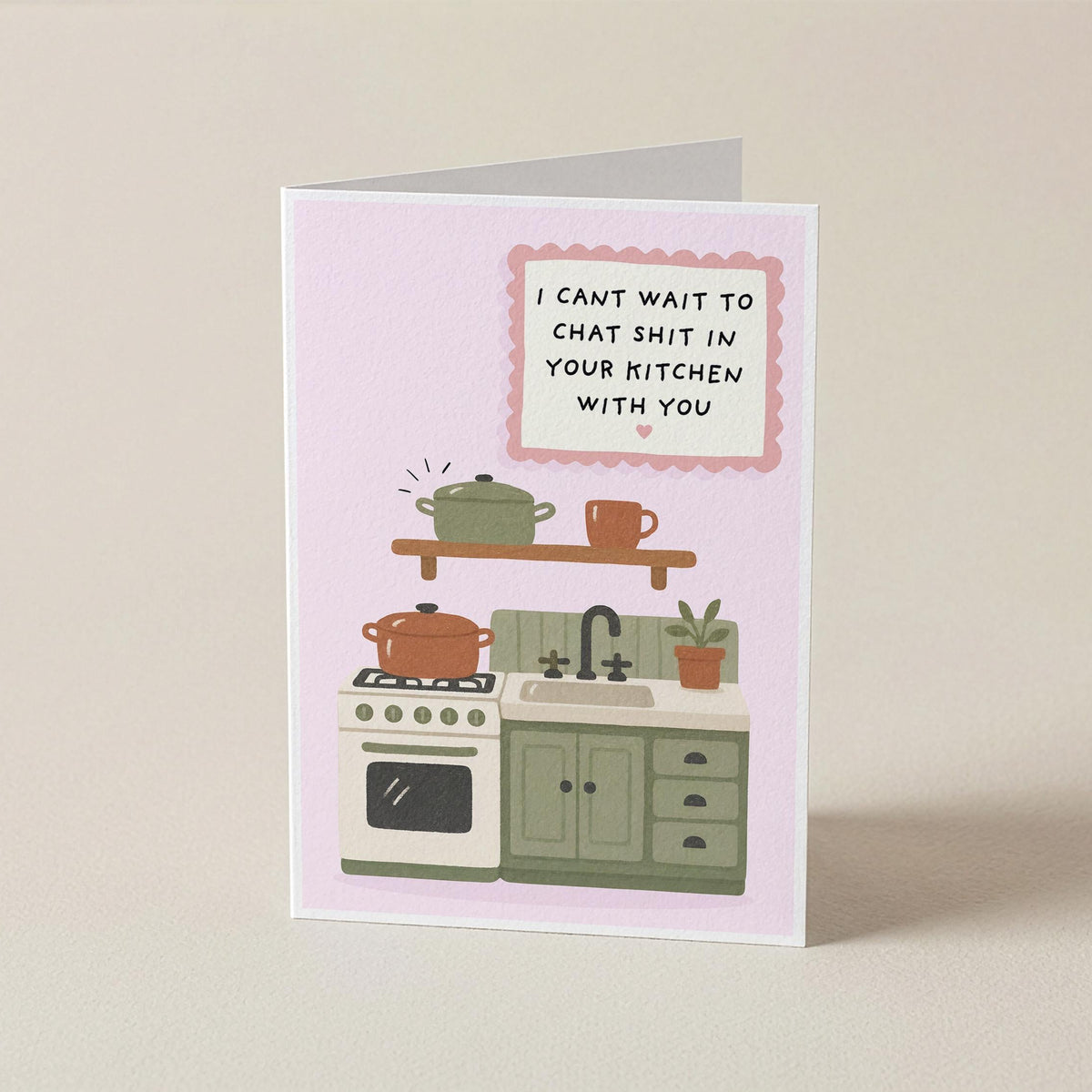 A folded greeting card standing upright on a light beige surface. The card has a pale pink background with a cartoon kitchen illustration and the text, 'I CAN'T WAIT TO CHAT SHIT IN YOUR KITCHEN WITH YOU.