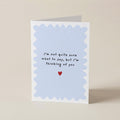 A folded greeting card standing upright on a light beige surface. The card has a white border with a pale blue scalloped inner frame and handwritten-style black text reading, 'I'm not quite sure what to say, but I'm thinking of you,' with a small red heart below.