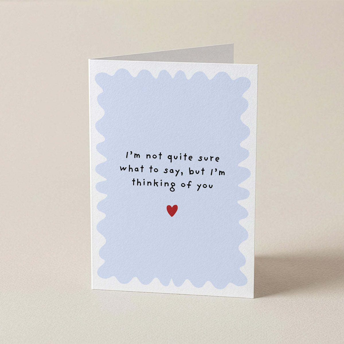 A folded greeting card standing upright on a light beige surface. The card has a white border with a pale blue scalloped inner frame and handwritten-style black text reading, 'I'm not quite sure what to say, but I'm thinking of you,' with a small red heart below.