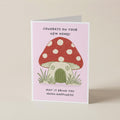 A folded greeting card standing upright on a light beige surface. The card has a pale pink background and features an illustration of a red mushroom house with white polka dots, green grass, and black text that reads, 'CONGRATS ON YOUR NEW HOME! MAY IT BRING YOU MUSH-HAPPINESS!'.