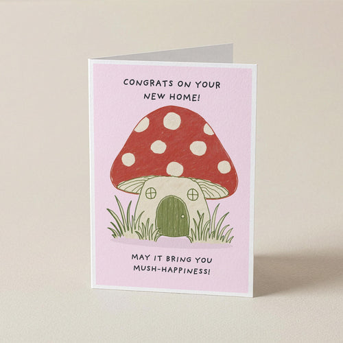 A folded greeting card standing upright on a light beige surface. The card has a pale pink background and features an illustration of a red mushroom house with white polka dots, green grass, and black text that reads, 'CONGRATS ON YOUR NEW HOME! MAY IT BRING YOU MUSH-HAPPINESS!'.