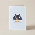 A folded greeting card standing upright on a light beige surface. The card features a cute illustration of a small black bat eating a two-tiered birthday cake against a pale blue rounded background.