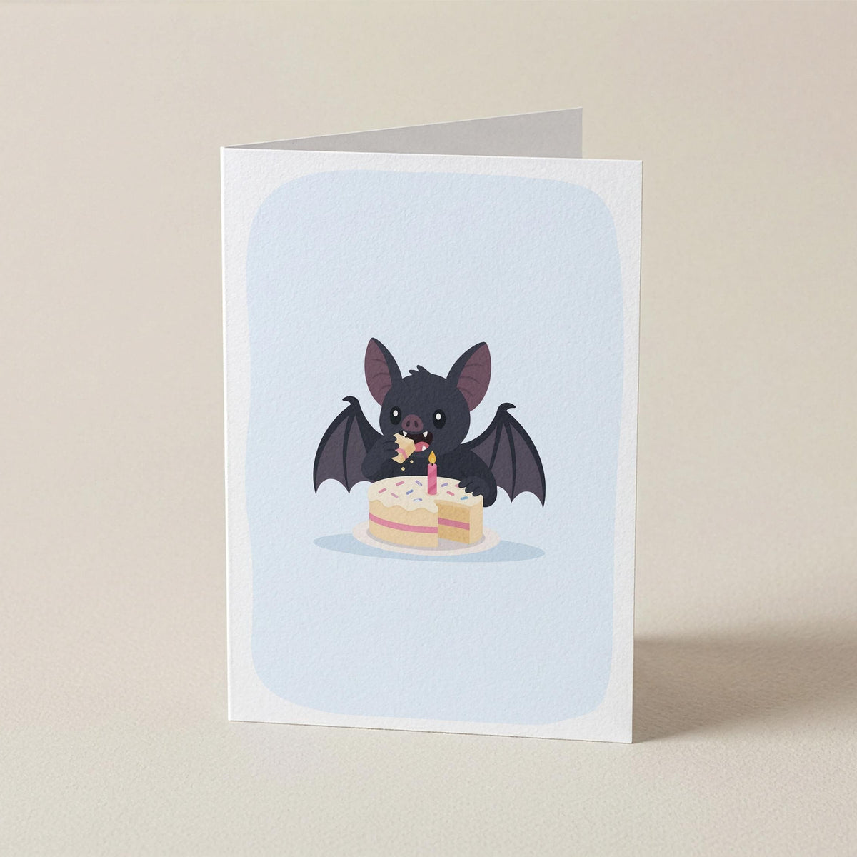 A folded greeting card standing upright on a light beige surface. The card features a cute illustration of a small black bat eating a two-tiered birthday cake against a pale blue rounded background.