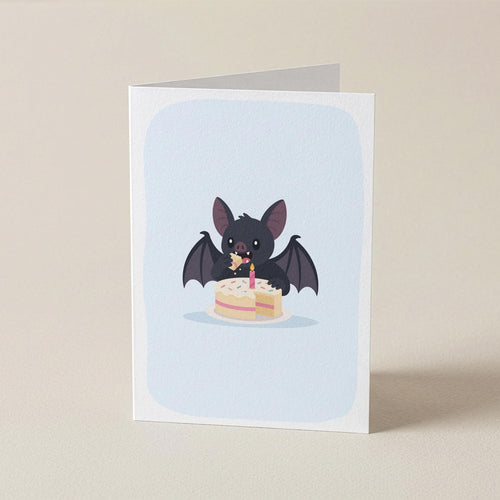 A folded greeting card standing upright on a light beige surface. The card features a cute illustration of a small black bat eating a two-tiered birthday cake against a pale blue rounded background.