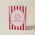 A greeting card with red and white vertical stripes and a central oval containing the text 'MILLIE, WILL YOU BE MY BRIDESMAID?' in red lettering, photographed on a plain light beige background.