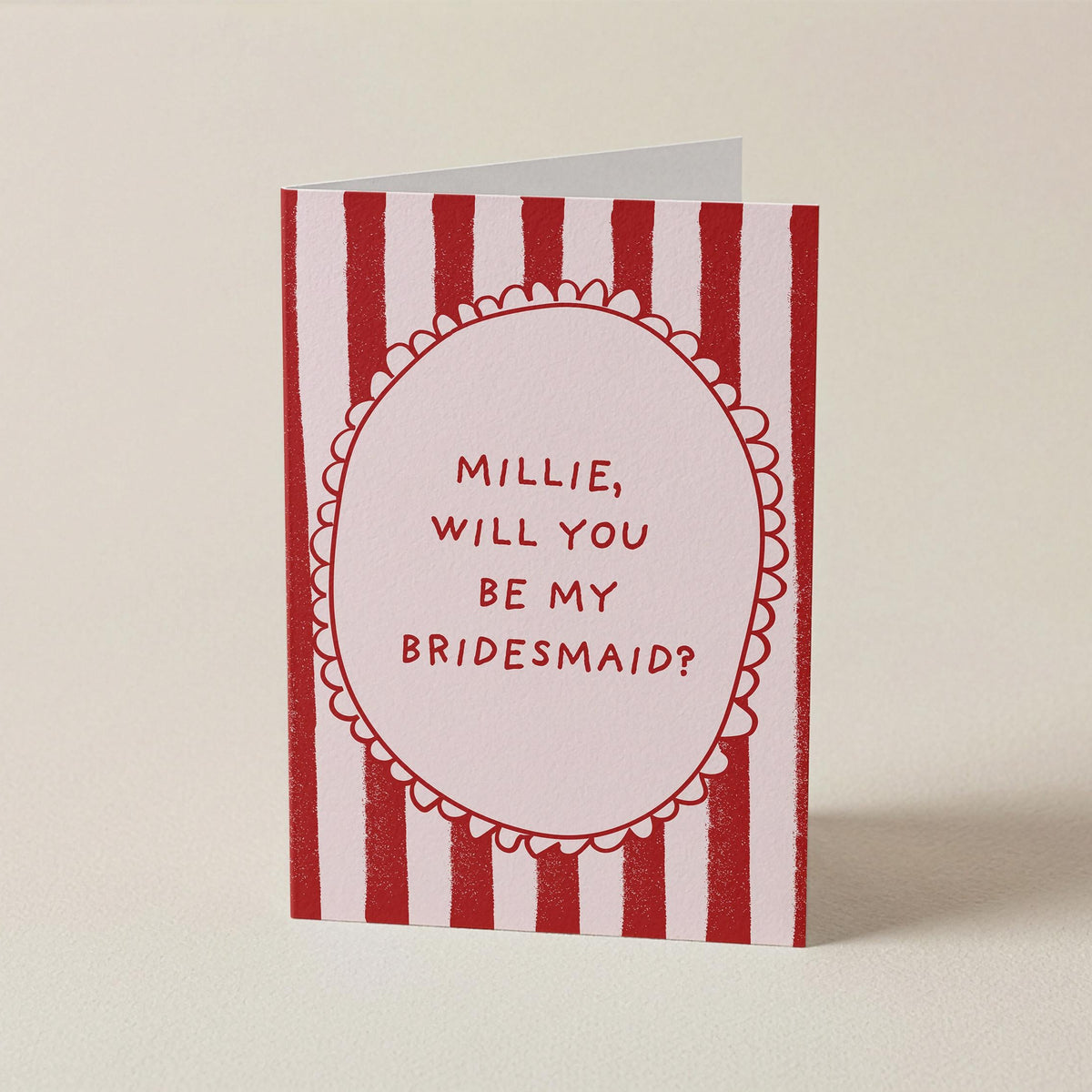A greeting card with red and white vertical stripes and a central oval containing the text 'MILLIE, WILL YOU BE MY BRIDESMAID?' in red lettering, photographed on a plain light beige background.