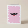 A folded greeting card standing upright on a light beige surface. The card has a white border and a pink scalloped center with the text 'HAPPY BIRTHDAY TO MY BESTIE' in black above a small red heart.