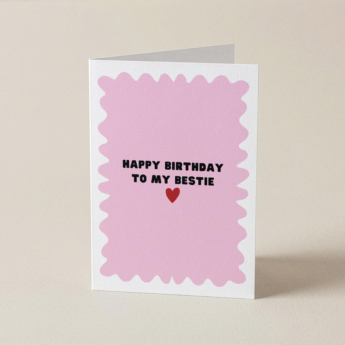 A folded greeting card standing upright on a light beige surface. The card has a white border and a pink scalloped center with the text 'HAPPY BIRTHDAY TO MY BESTIE' in black above a small red heart.