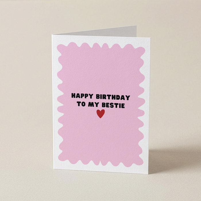 A folded greeting card standing upright on a light beige surface. The card has a white border and a pink scalloped center with the text 'HAPPY BIRTHDAY TO MY BESTIE' in black above a small red heart.
