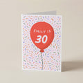 A folded greeting card standing upright on a beige surface. The card features a white background covered in small multicolored confetti-like specks, with a large red balloon graphic in the center containing the text 'EMILY IS 30' in white.