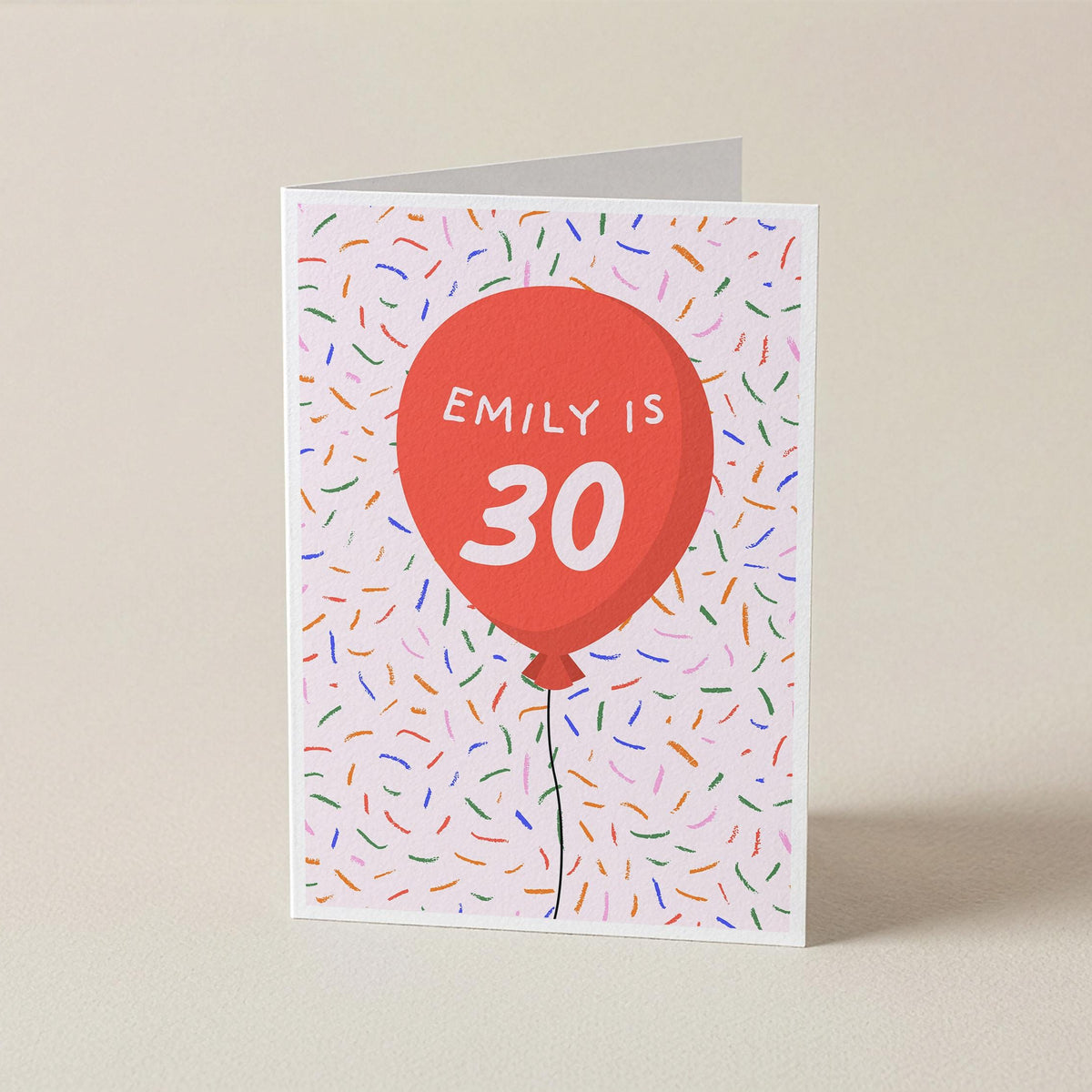 A folded greeting card standing upright on a beige surface. The card features a white background covered in small multicolored confetti-like specks, with a large red balloon graphic in the center containing the text 'EMILY IS 30' in white.