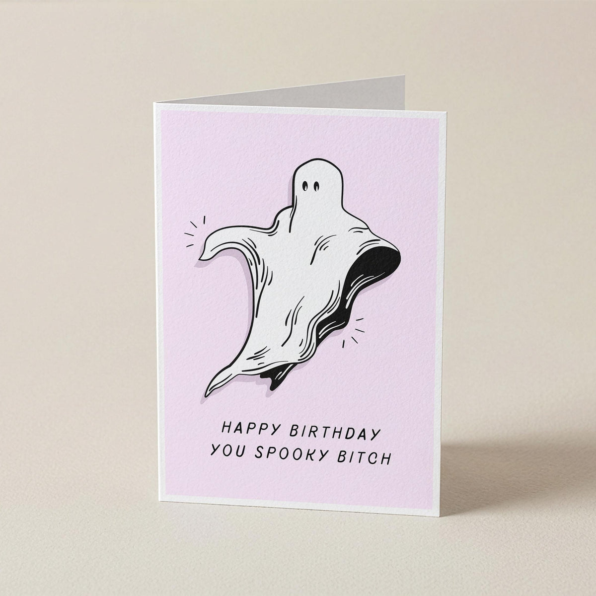 A folded greeting card standing upright on a light beige surface. The card has a pale pink front with a black-and-white ghost illustration and the text 'HAPPY BIRTHDAY YOU SPOOKY BITCH'.