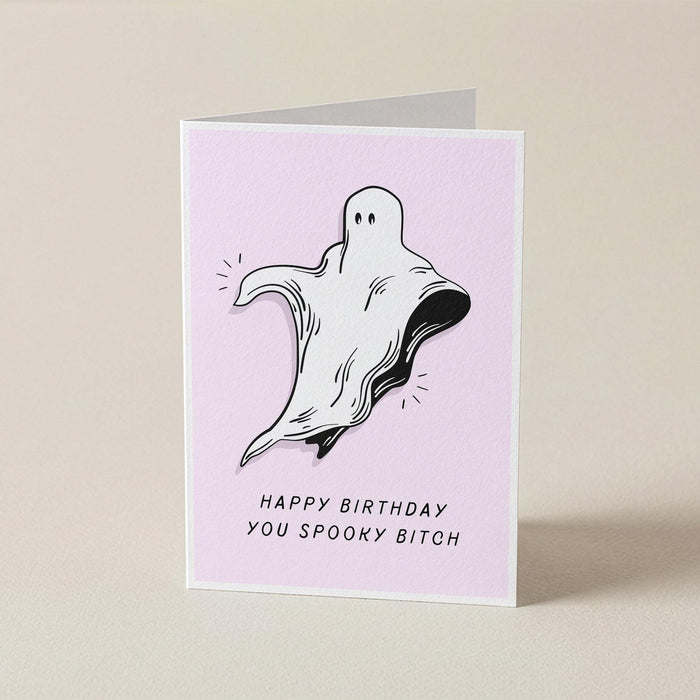 A folded greeting card standing upright on a light beige surface. The card has a pale pink front with a black-and-white ghost illustration and the text 'HAPPY BIRTHDAY YOU SPOOKY BITCH'.