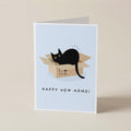 A folded greeting card standing upright on a beige surface. The card features a light blue background with an illustration of a black cat sitting inside a cardboard box, above the text 'HAPPY NEW HOME!'.