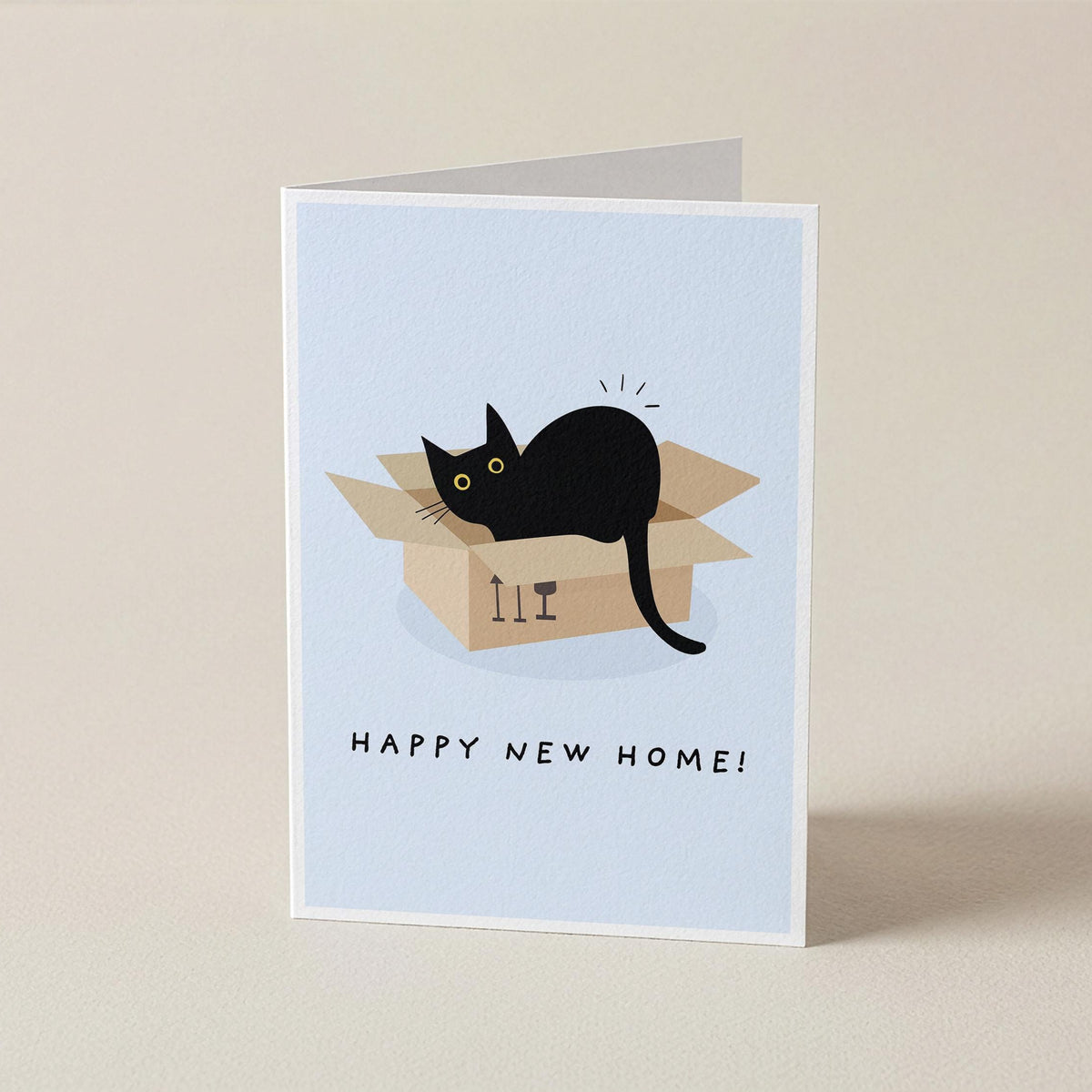 A folded greeting card standing upright on a beige surface. The card features a light blue background with an illustration of a black cat sitting inside a cardboard box, above the text 'HAPPY NEW HOME!'.