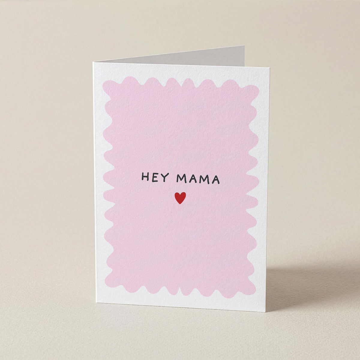 A folded greeting card standing upright on a light beige surface. The front of the card has a white border with a scalloped pink inner design and the words 'HEY MAMA' in black above a small red heart.