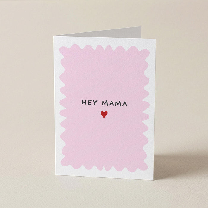 A folded greeting card standing upright on a light beige surface. The front of the card has a white border with a scalloped pink inner design and the words 'HEY MAMA' in black above a small red heart.