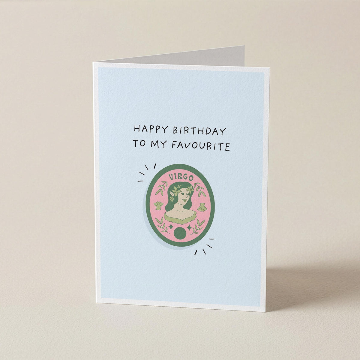 A folded greeting card standing upright on a light beige surface. The card has a pale blue front with the text 'HAPPY BIRTHDAY TO MY FAVOURITE' above a circular illustration featuring a woman's profile and the word 'VIRGO'.