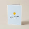 A folded greeting card standing upright on a warm beige surface. The front of the card is pale blue with a small yellow sun illustration and the text 'SENDING YOU SOME SUNSHINE' in black.