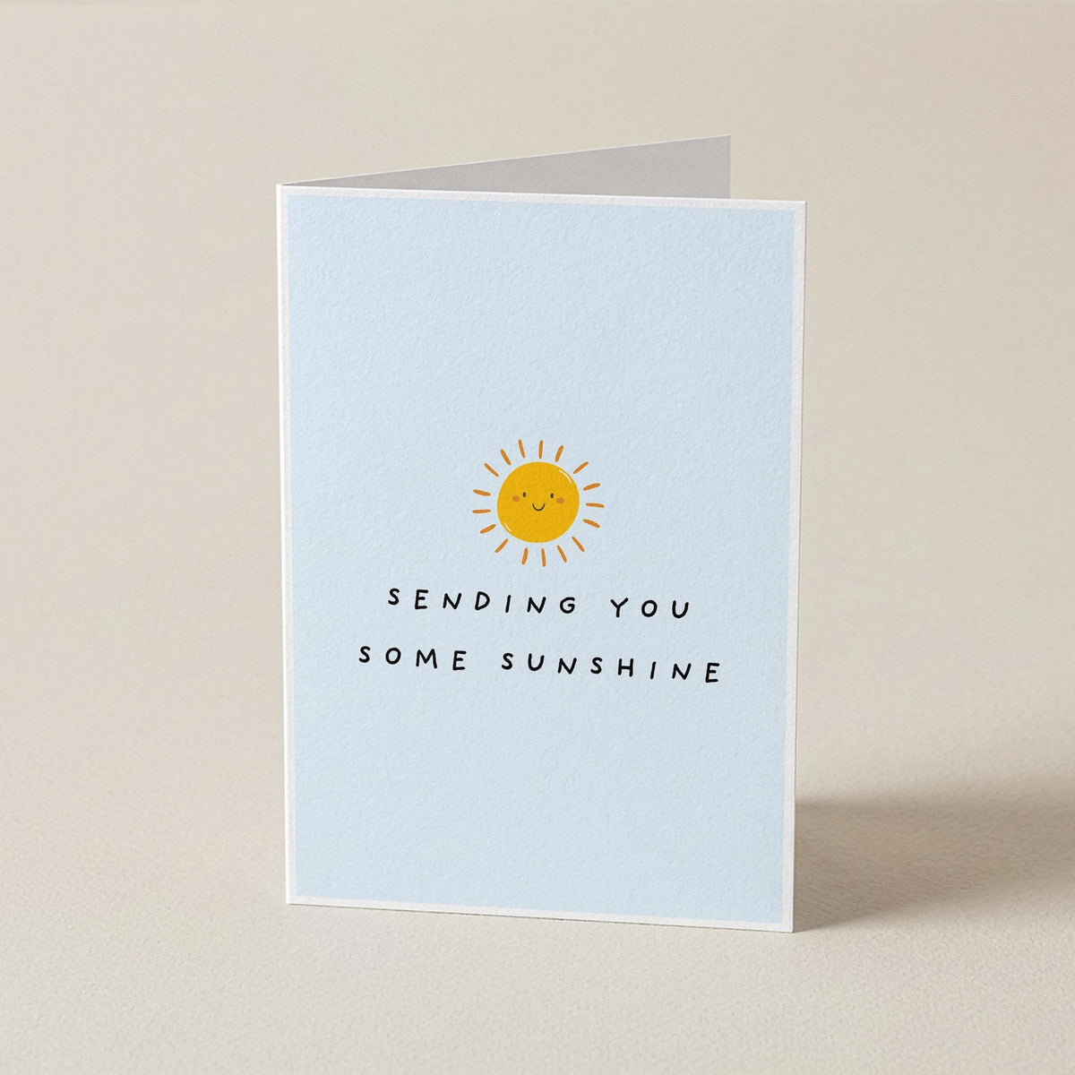 A folded greeting card standing upright on a warm beige surface. The front of the card is pale blue with a small yellow sun illustration and the text 'SENDING YOU SOME SUNSHINE' in black.