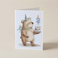A folded greeting card standing upright on a light beige surface. The card features a watercolor illustration of a brown teddy bear wearing a party hat and bow tie, holding a small birthday cake with a blue candle shaped like the number 2.