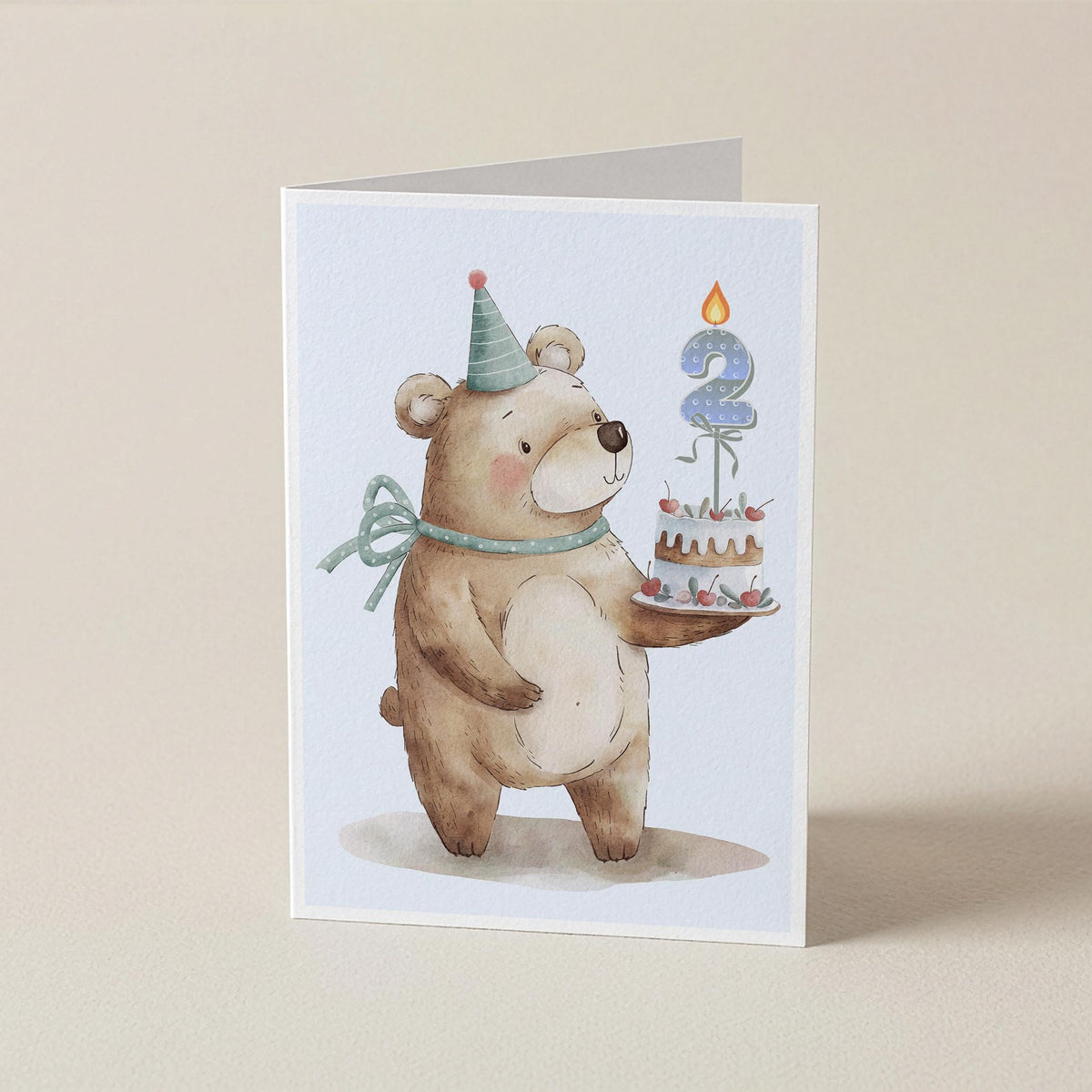 A folded greeting card standing upright on a light beige surface. The card features a watercolor illustration of a brown teddy bear wearing a party hat and bow tie, holding a small birthday cake with a blue candle shaped like the number 2.