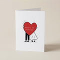 A white greeting card standing upright on a light beige surface, featuring a simple illustration of a person in black pants and a white dress holding a large red heart with the words 'HERE'S TO FOREVER' inside.