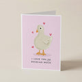 A folded greeting card standing upright on a light beige surface. The card has a pale pink background with a cartoon duck in white and orange, small pink hearts, and the text 'I LOVE YOU SO DUCKING MUCH' at the bottom.