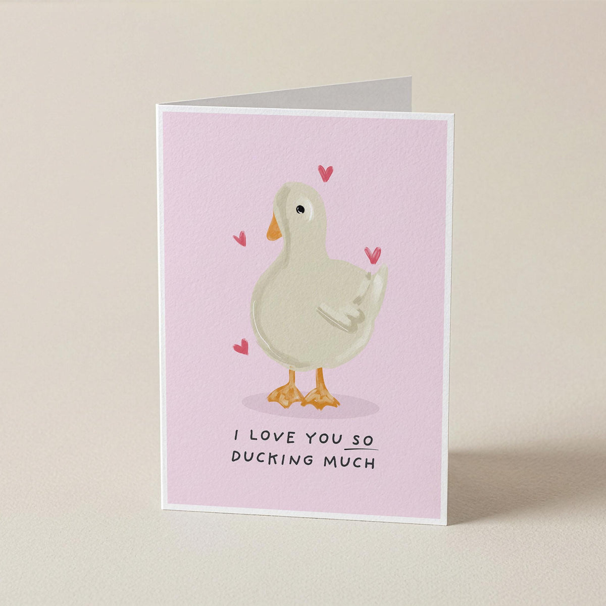 A folded greeting card standing upright on a light beige surface. The card has a pale pink background with a cartoon duck in white and orange, small pink hearts, and the text 'I LOVE YOU SO DUCKING MUCH' at the bottom.