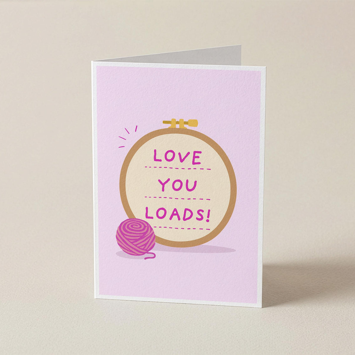 A folded greeting card standing upright on a light beige surface. The card has a pale pink background with a central embroidery hoop illustration containing the phrase 'LOVE YOU LOADS!' in pink text, plus a pink yarn ball graphic near the bottom left.