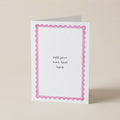 A white greeting card with a pink scalloped border and the text 'add your own text here' printed in the center, standing upright on a light beige background.