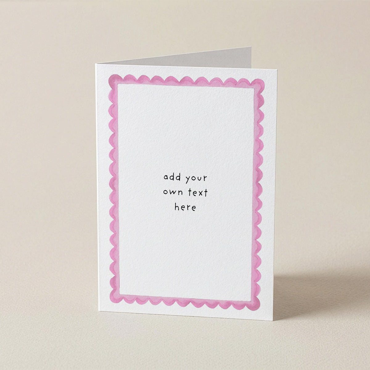 A white greeting card with a pink scalloped border and the text 'add your own text here' printed in the center, standing upright on a light beige background.