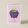 A folded greeting card standing upright on a light beige surface. The card has a pink background with a cartoon purple cauldron filled with green potion and the text 'witching you a happy birthday!'.