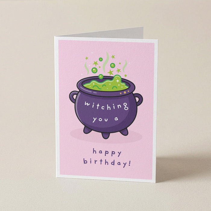A folded greeting card standing upright on a light beige surface. The card has a pink background with a cartoon purple cauldron filled with green potion and the text 'witching you a happy birthday!'.