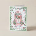 A personalized Christmas greeting card with a white background, featuring a red illustrated door decorated with a green wreath and red bow, surrounded by a border of green holly leaves and red berries. The text 'STACEY & HARRY' is printed at the bottom.