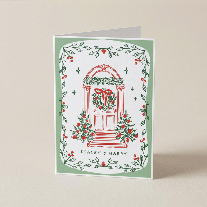 A personalized Christmas greeting card with a white background, featuring a red illustrated door decorated with a green wreath and red bow, surrounded by a border of green holly leaves and red berries. The text 'STACEY & HARRY' is printed at the bottom.